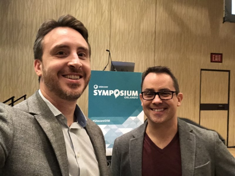 Mark Stiles and Dan Murphy stand together smiling with a sign for Sitecore Symposium in the background.